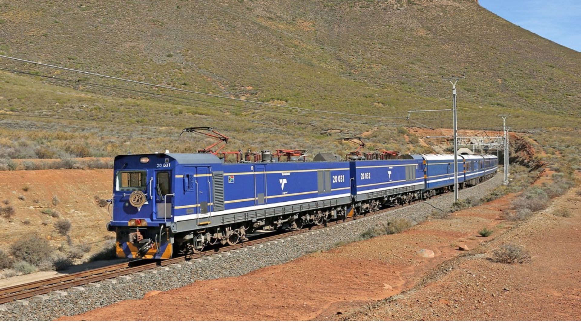 A blue electric train with several passenger cars travels through a dry, hilly landscape dotted with bush and sparse vegetation, offering the perfect travel getaway beneath a clear sky and low mountains.