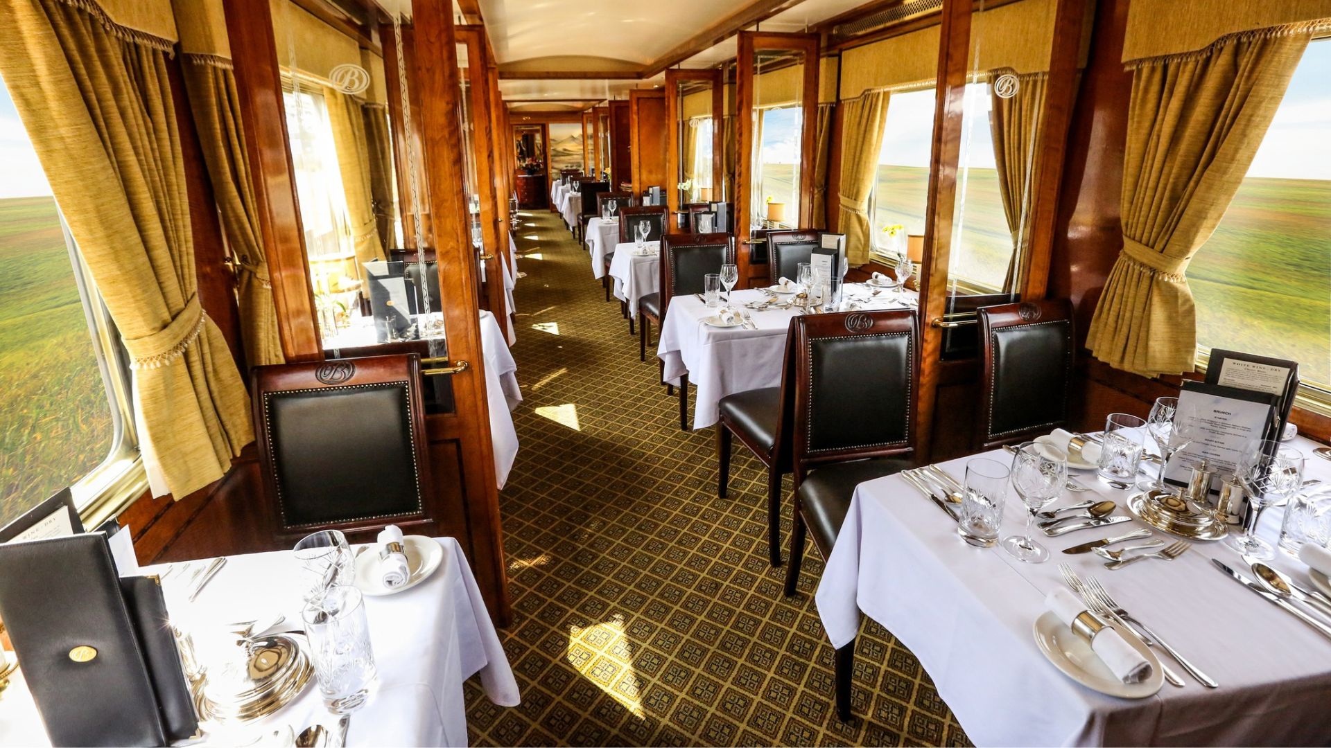 Elegant train dining car with tables set for a meal, perfect for a scenic getaway. White tablecloths, glassware, and menus rest on polished wood with gold accents, while large windows reveal bush-lined green fields in bright daylight.