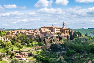 Hilltop Italian village with terracotta-roofed buildings, a prominent church tower, and lush green trees and bush, surrounded by rolling hills and fields—a perfect travel getaway under a partly cloudy sky.