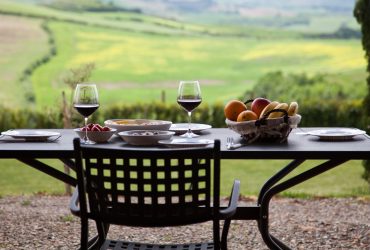 A set table for two outdoors, featuring two glasses of red wine, bowls of food, and a basket of fresh fruit, perfect for a peaceful getaway with a scenic view of green fields and hills in the background.