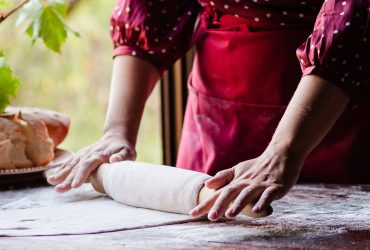 A person in a red apron and polka-dot shirt rolls out dough on a floured wooden surface, evoking the cozy charm of a travel getaway, with a loaf of bread and green leaves visible in the background by a window.