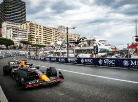 A Formula 1 car races along a street circuit by the waterfront, with tall buildings, yachts, and spectators in the background under a cloudy sky. MSC Cruises banners line the track, capturing the thrill of travel and a high-speed getaway.