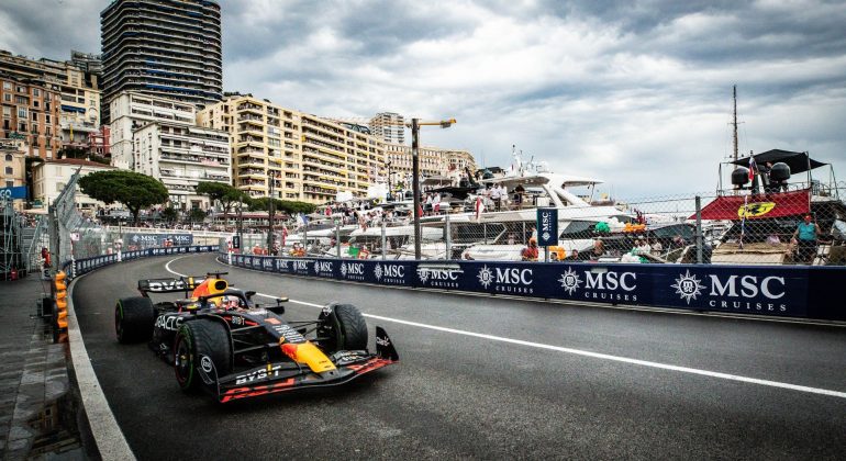 A Formula 1 car races along a street circuit by the waterfront, with tall buildings, yachts, and spectators in the background under a cloudy sky. MSC Cruises banners line the track, capturing the thrill of travel and a high-speed getaway.