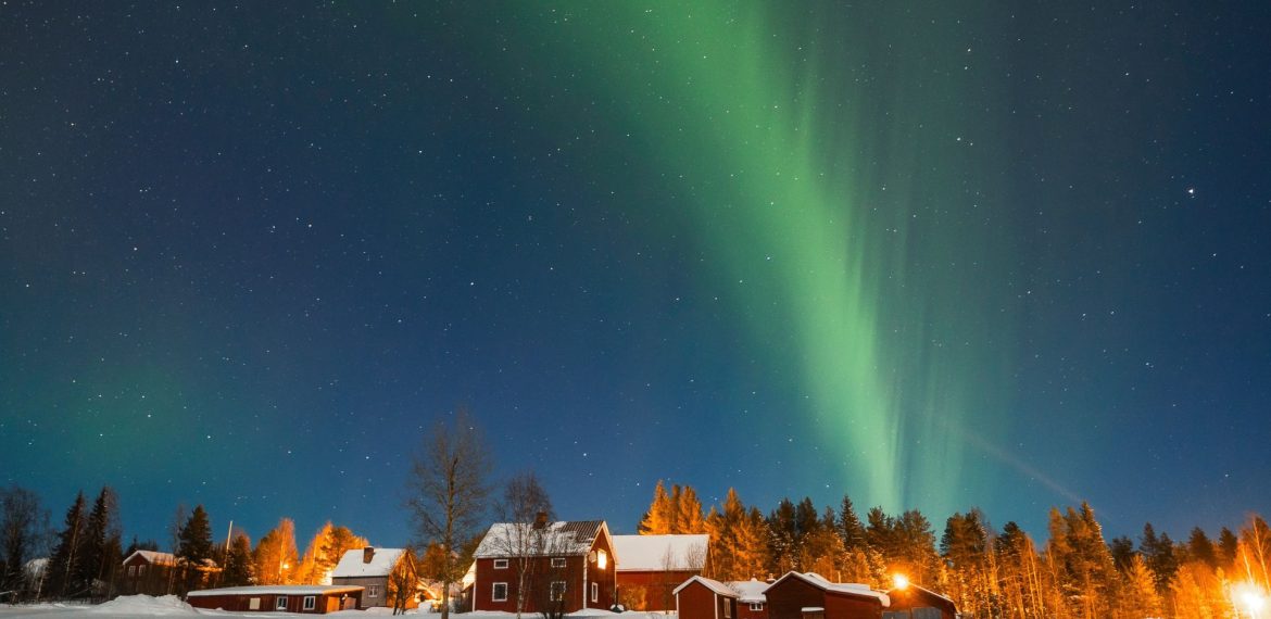 Green aurora borealis lights up the night sky above red wooden houses and snow-covered ground, creating a magical travel getaway with trees, bush, and twinkling stars in the background.