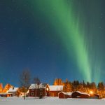 Green aurora borealis lights up the night sky above red wooden houses and snow-covered ground, creating a magical travel getaway with trees, bush, and twinkling stars in the background.