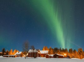 Green aurora borealis lights up the night sky above red wooden houses and snow-covered ground, creating a magical travel getaway with trees, bush, and twinkling stars in the background.