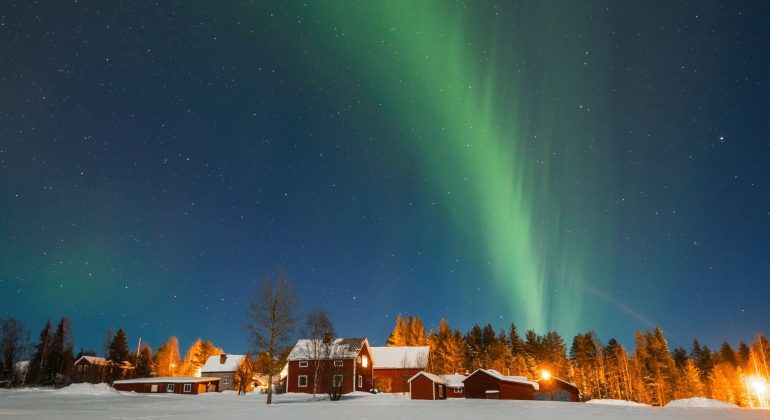 Green aurora borealis lights up the night sky above red wooden houses and snow-covered ground, creating a magical travel getaway with trees, bush, and twinkling stars in the background.