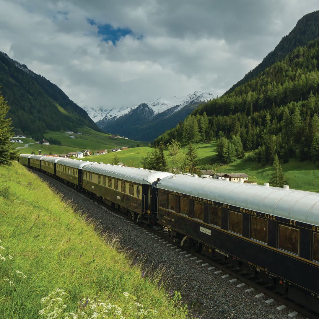 A vintage luxury train offers the perfect travel getaway as it winds through a lush green valley, bush-lined hills, and snow-capped mountains under a partly cloudy sky. Small houses dot the distant landscape.