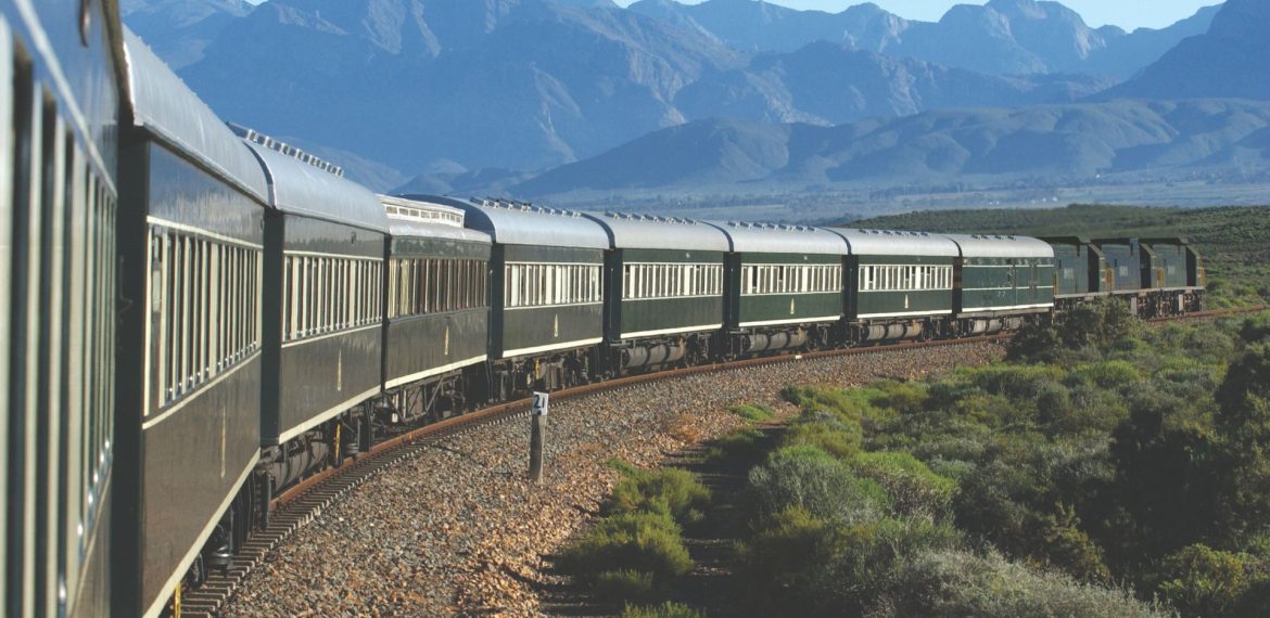 A long, green passenger train moves along a curved track through bush and lush landscape, with mountains in the background under a clear blue sky—the perfect scene for a memorable travel getaway.