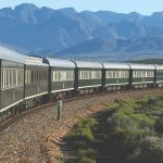 A long, green passenger train moves along a curved track through bush and lush landscape, with mountains in the background under a clear blue sky—the perfect scene for a memorable travel getaway.