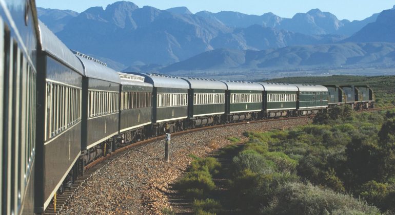 A long, green passenger train moves along a curved track through bush and lush landscape, with mountains in the background under a clear blue sky—the perfect scene for a memorable travel getaway.