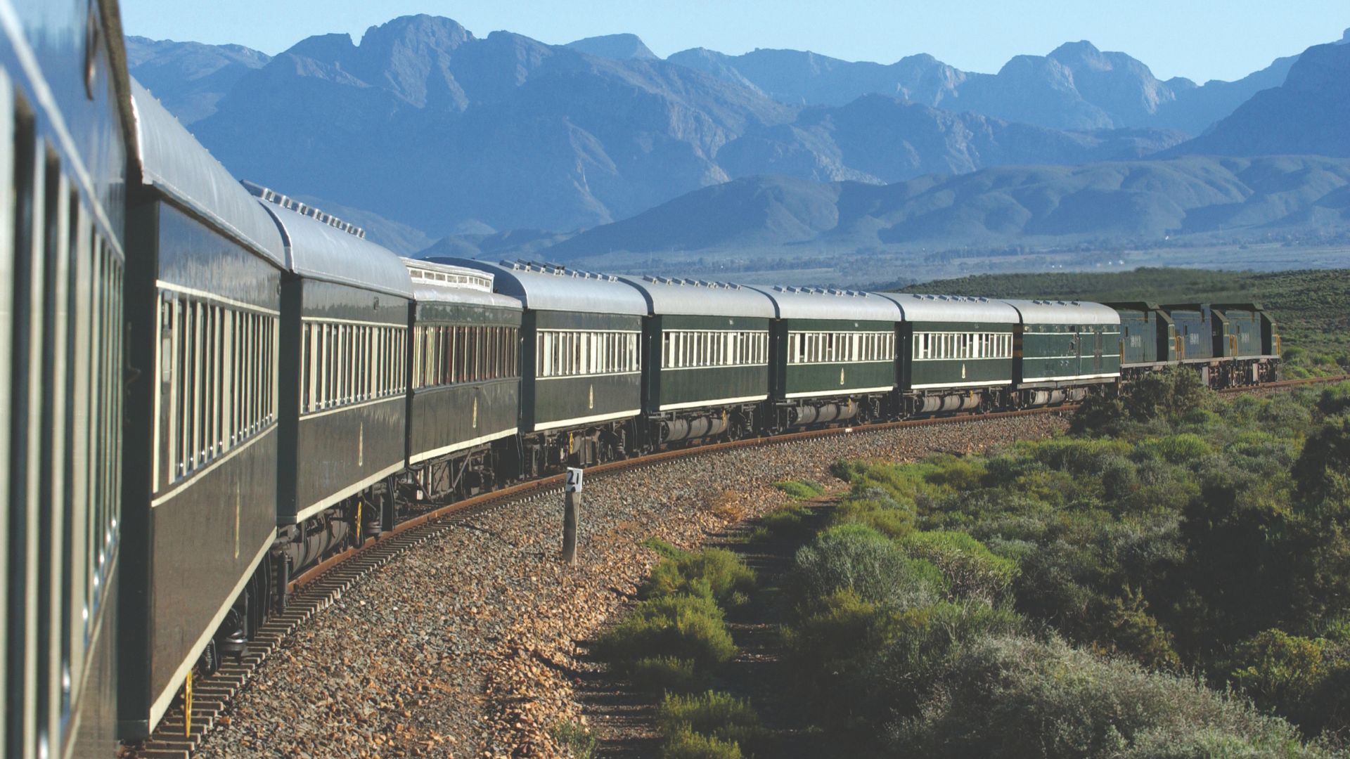 A long, green passenger train moves along a curved track through bush and lush landscape, with mountains in the background under a clear blue sky—the perfect scene for a memorable travel getaway.