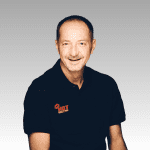 Patrick, a middle-aged man with short brown hair and a mustache, smiles at the camera in a black polo shirt featuring the HOT 102.7 FM logo, set against a light gray gradient background.