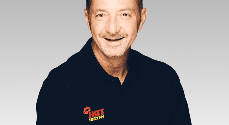 Patrick, a middle-aged man with short brown hair and a mustache, smiles at the camera in a black polo shirt featuring the HOT 102.7 FM logo, set against a light gray gradient background.