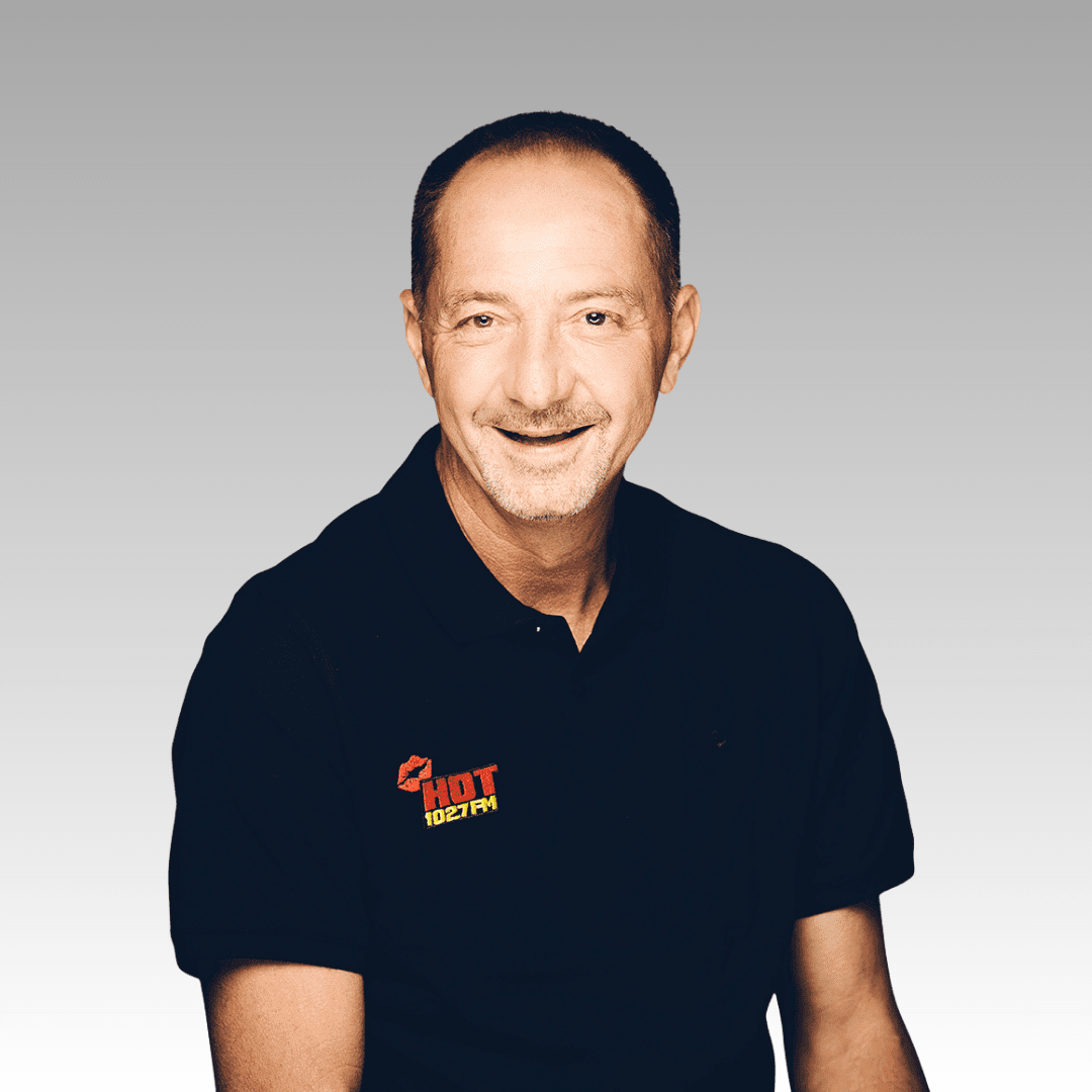 Patrick, a middle-aged man with short brown hair and a mustache, smiles at the camera in a black polo shirt featuring the HOT 102.7 FM logo, set against a light gray gradient background.
