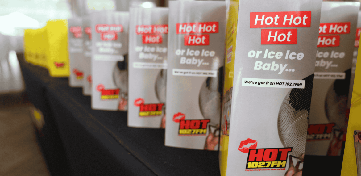 Several table tents featuring the HOT 102.7 FM logo and playful phrases like "Hot Hot Hot" or "Ice Ice Baby…" are lined up across a black-clothed table, setting the stage for a sizzling hot 1027 breakfast event.