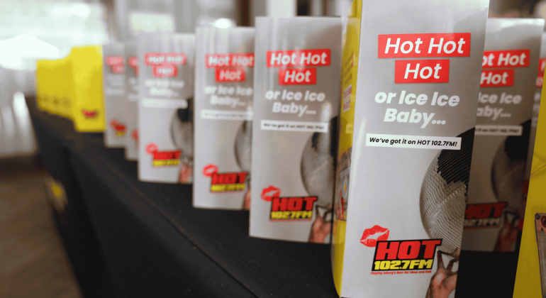 Several table tents featuring the HOT 102.7 FM logo and playful phrases like "Hot Hot Hot" or "Ice Ice Baby…" are lined up across a black-clothed table, setting the stage for a sizzling hot 1027 breakfast event.