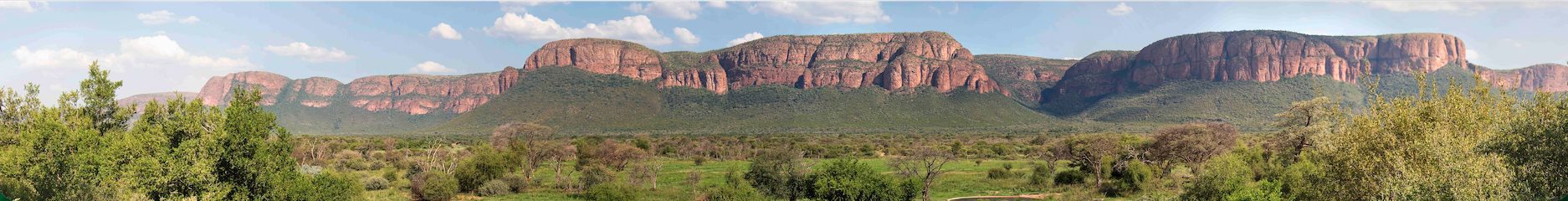 Panoramic view of a green savanna with lush trees in the foreground and a large, red rocky mountain range under a blue sky with scattered clouds—perfect scenery for a memorable Matric celebration.