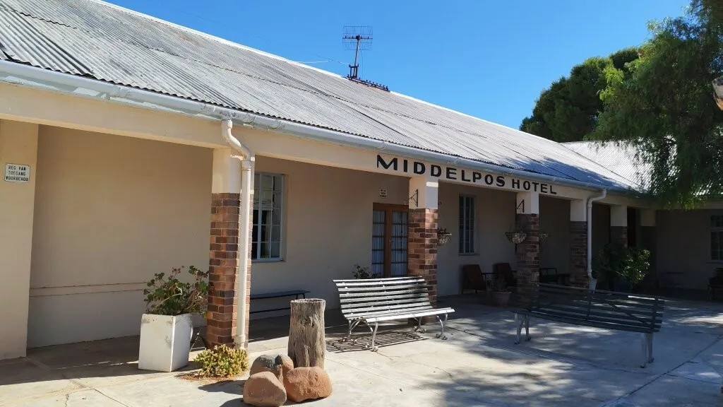 The front of Middelpos Hotel with a corrugated metal roof, brick pillars, benches, potted plants, and a sunny, clear sky—an oasis far from the bustle of Johannesburg water concerns.