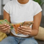 A person in a white t-shirt and jeans sits on a couch, holding and counting various colorful banknotes. Standard Bank logos are visible on some of the notes, with yellow and green pillows in the background.