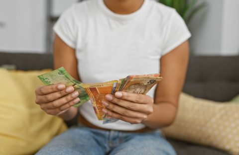 A person in a white t-shirt and jeans sits on a couch, holding and counting various colorful banknotes. Standard Bank logos are visible on some of the notes, with yellow and green pillows in the background.