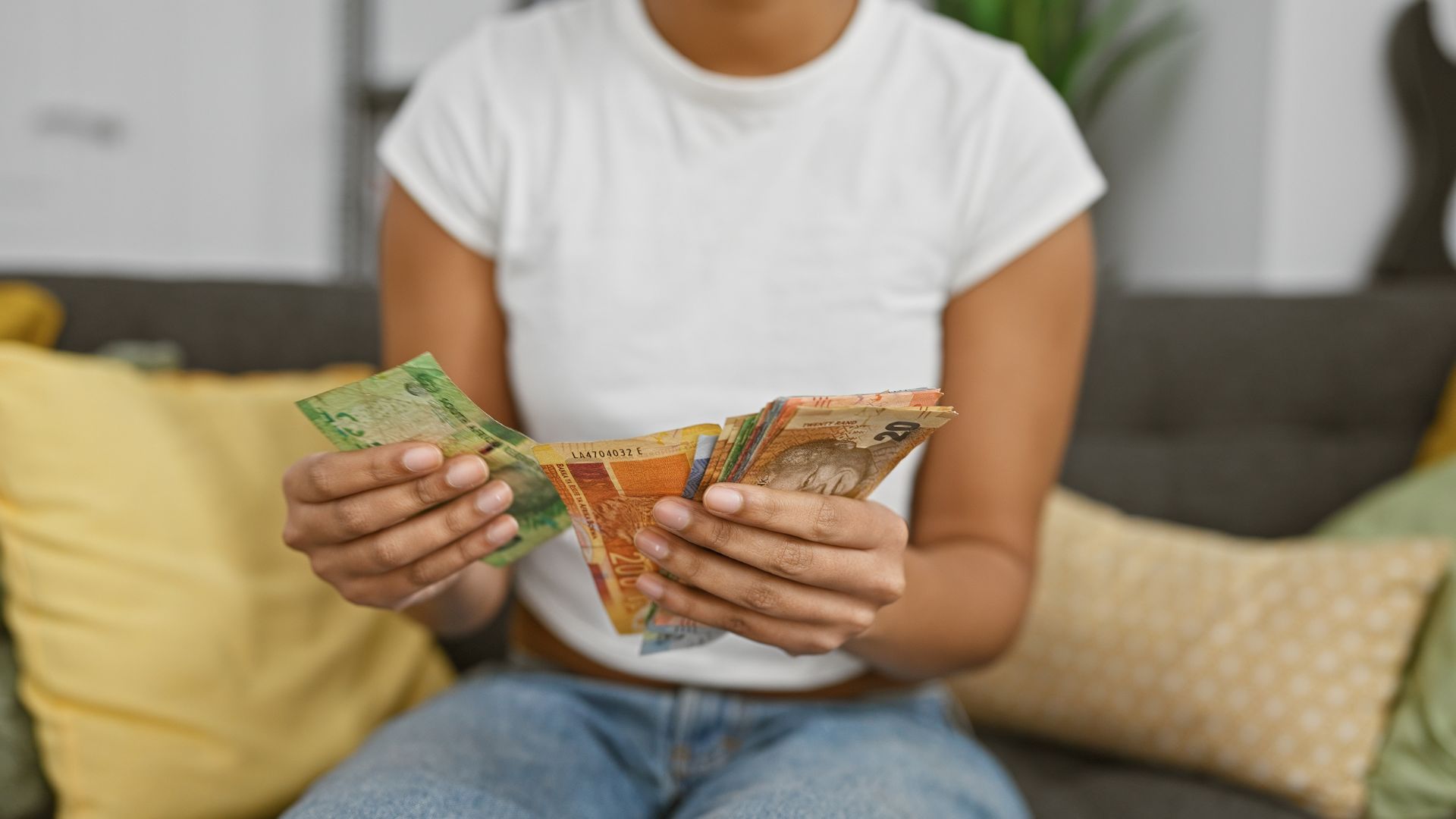 A person in a white t-shirt and jeans sits on a couch, holding and counting various colorful banknotes. Standard Bank logos are visible on some of the notes, with yellow and green pillows in the background.