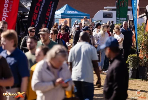 A crowd of people walk and gather among vendor tents and banners at the Kingsmead Book Fair on a sunny day. Some interact with each other, while others look at their phones or stroll through the lively outdoor scene.