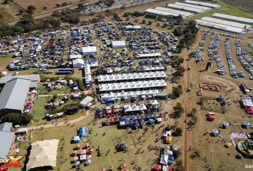 Aerial view of a large outdoor festival, reminiscent of the Kingsmead Book Fair, with rows of white tents, numerous booths, people walking, parked cars, and open fields; logo Outdoor X is visible in the bottom left corner.
