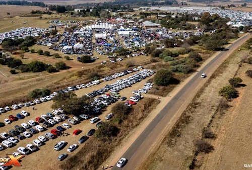 Aerial view of Kingsmead Book Fair, a large outdoor event with numerous white tents and crowds, surrounded by fields. Many cars are parked in rows on dry grass near a paved road. Outdoor X and Gary Media logos are visible.