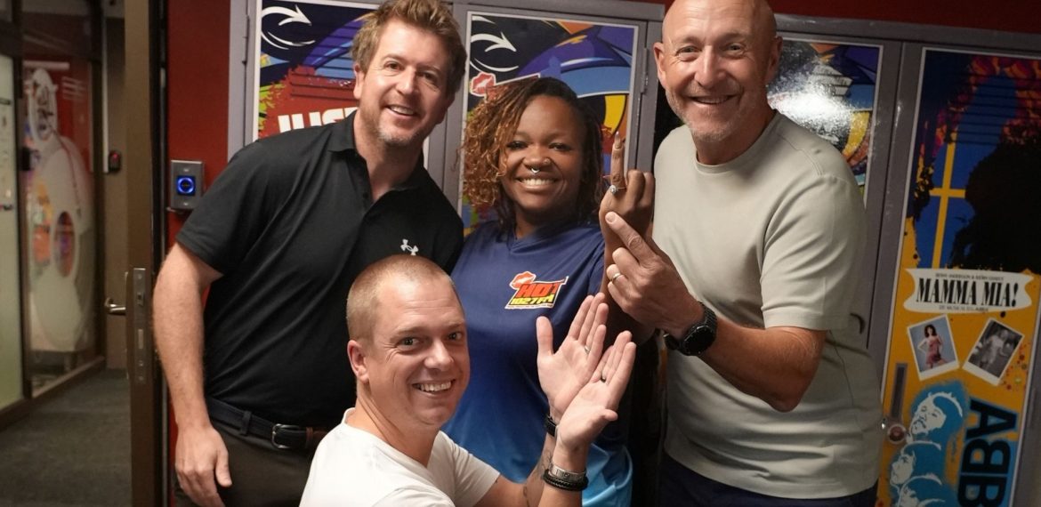 Four smiling adults pose together indoors in front of colorful lockers, exuding cheerful and friendly vibes—like a happy crew at a Hot 1027 breakfast, with one person kneeling in front while the others stand closely behind.