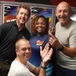 Four smiling adults pose together indoors in front of colorful lockers, exuding cheerful and friendly vibes—like a happy crew at a Hot 1027 breakfast, with one person kneeling in front while the others stand closely behind.