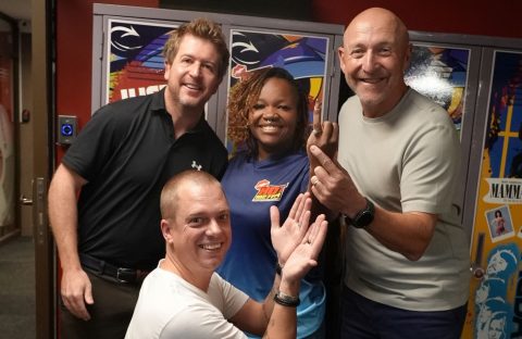 Four smiling adults pose together indoors in front of colorful lockers, exuding cheerful and friendly vibes—like a happy crew at a Hot 1027 breakfast, with one person kneeling in front while the others stand closely behind.
