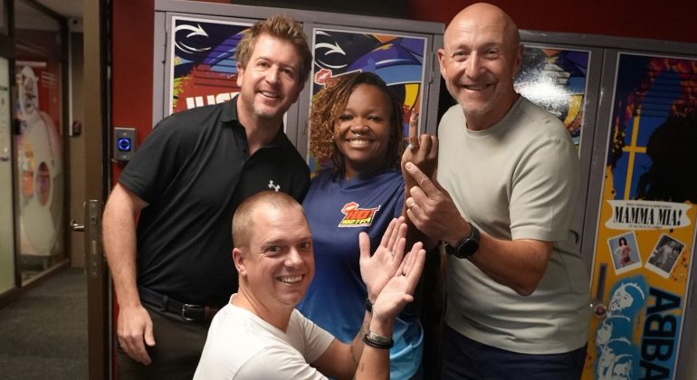 Four smiling adults pose together indoors in front of colorful lockers, exuding cheerful and friendly vibes—like a happy crew at a Hot 1027 breakfast, with one person kneeling in front while the others stand closely behind.