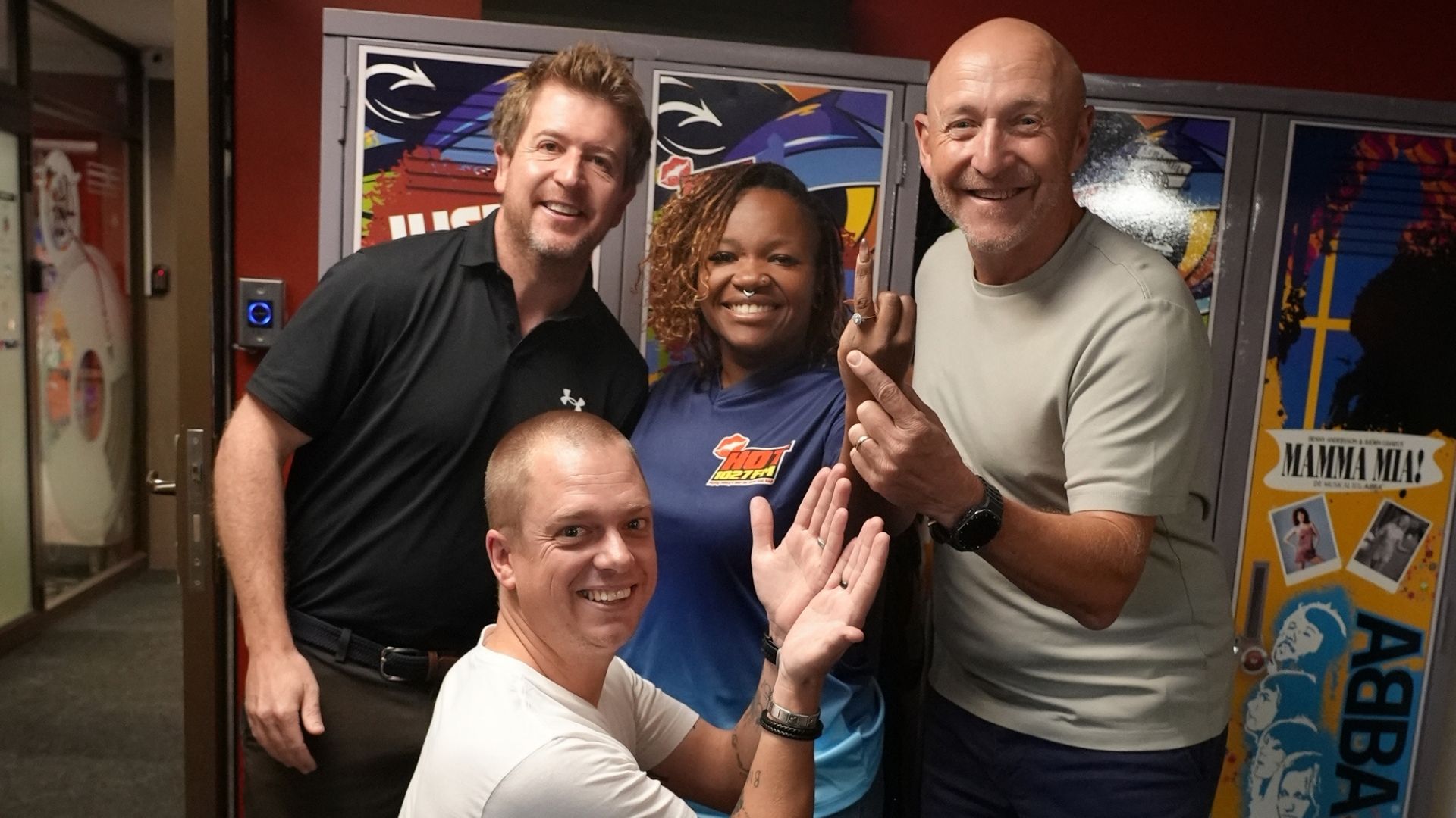 Four smiling adults pose together indoors in front of colorful lockers, exuding cheerful and friendly vibes—like a happy crew at a Hot 1027 breakfast, with one person kneeling in front while the others stand closely behind.