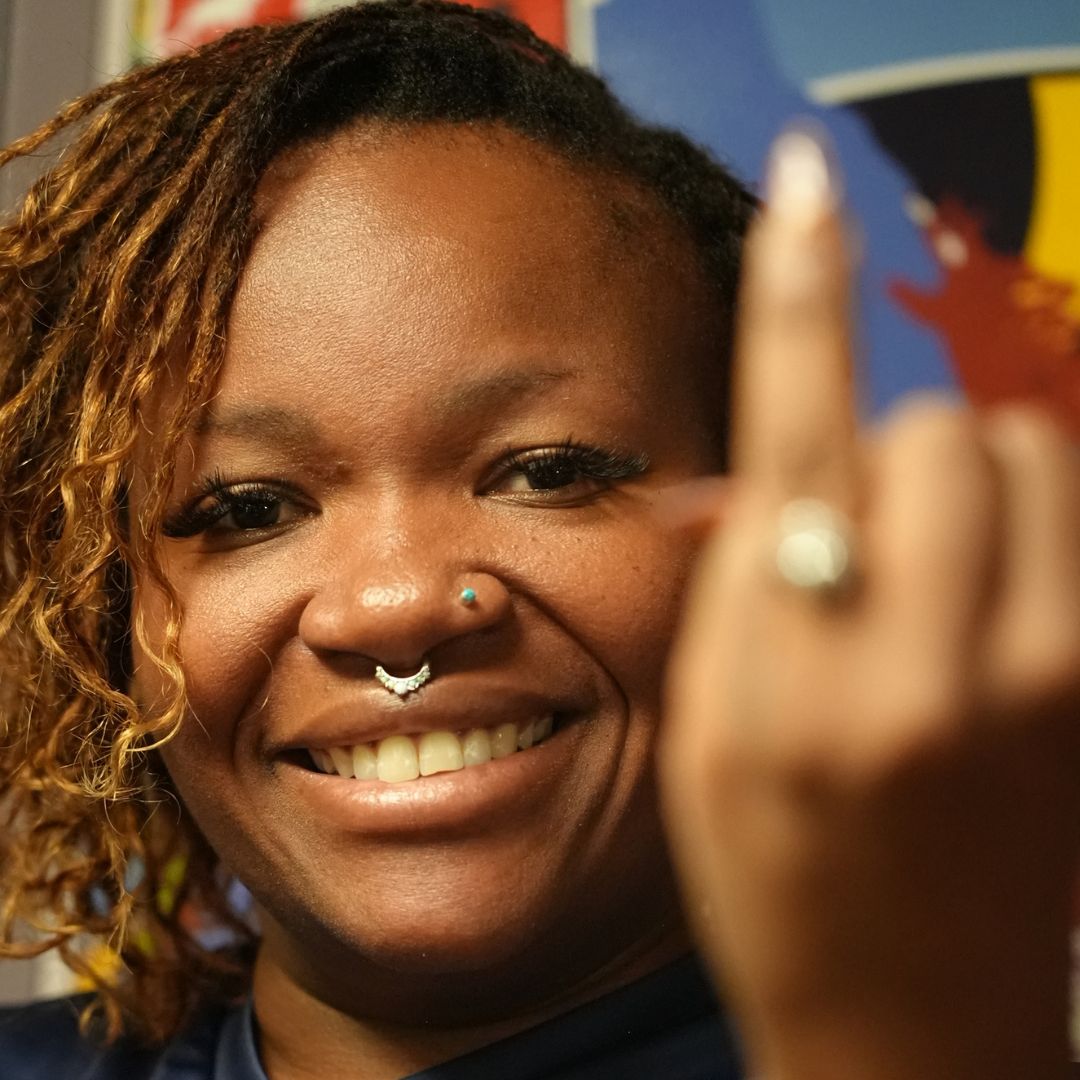 A smiling person with a nose ring and curly hair holds up their middle finger towards the camera, partially out of focus, against a colorful backdrop—capturing the bold energy of a Hot 1027 Breakfast vibe.