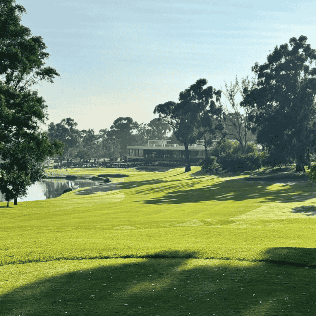 A lush golf course with neatly mowed grass, bordered by tall trees. A calm water feature offers a sense of fuel relief, while the distant clubhouse stands under a clear blue sky and tree shadows stretch across the grass.
