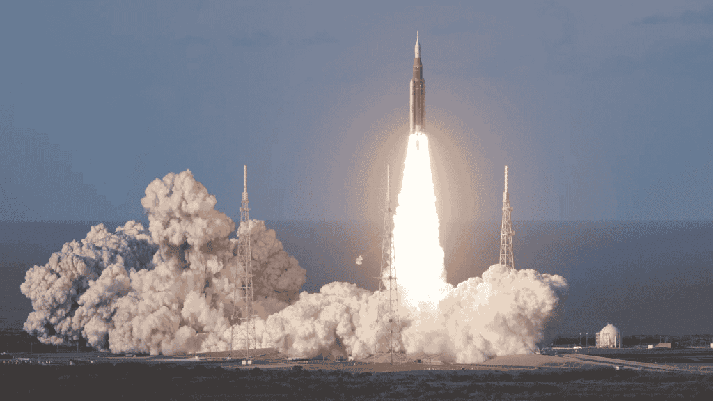 A rocket launches into the sky, surrounded by clouds of smoke and bright flames at its base, reminiscent of the energy at a hot 1027 breakfast, with support towers and a clear sky in the background.