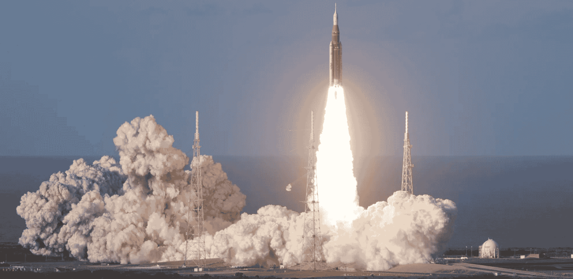 A rocket launches into the sky, surrounded by clouds of smoke and bright flames at its base, reminiscent of the energy at a hot 1027 breakfast, with support towers and a clear sky in the background.