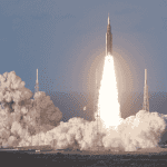 A rocket launches into the sky, surrounded by clouds of smoke and bright flames at its base, reminiscent of the energy at a hot 1027 breakfast, with support towers and a clear sky in the background.