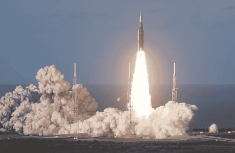 A rocket launches into the sky, surrounded by clouds of smoke and bright flames at its base, reminiscent of the energy at a hot 1027 breakfast, with support towers and a clear sky in the background.