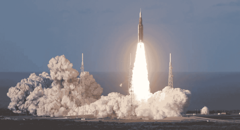 A rocket launches into the sky, surrounded by clouds of smoke and bright flames at its base, reminiscent of the energy at a hot 1027 breakfast, with support towers and a clear sky in the background.