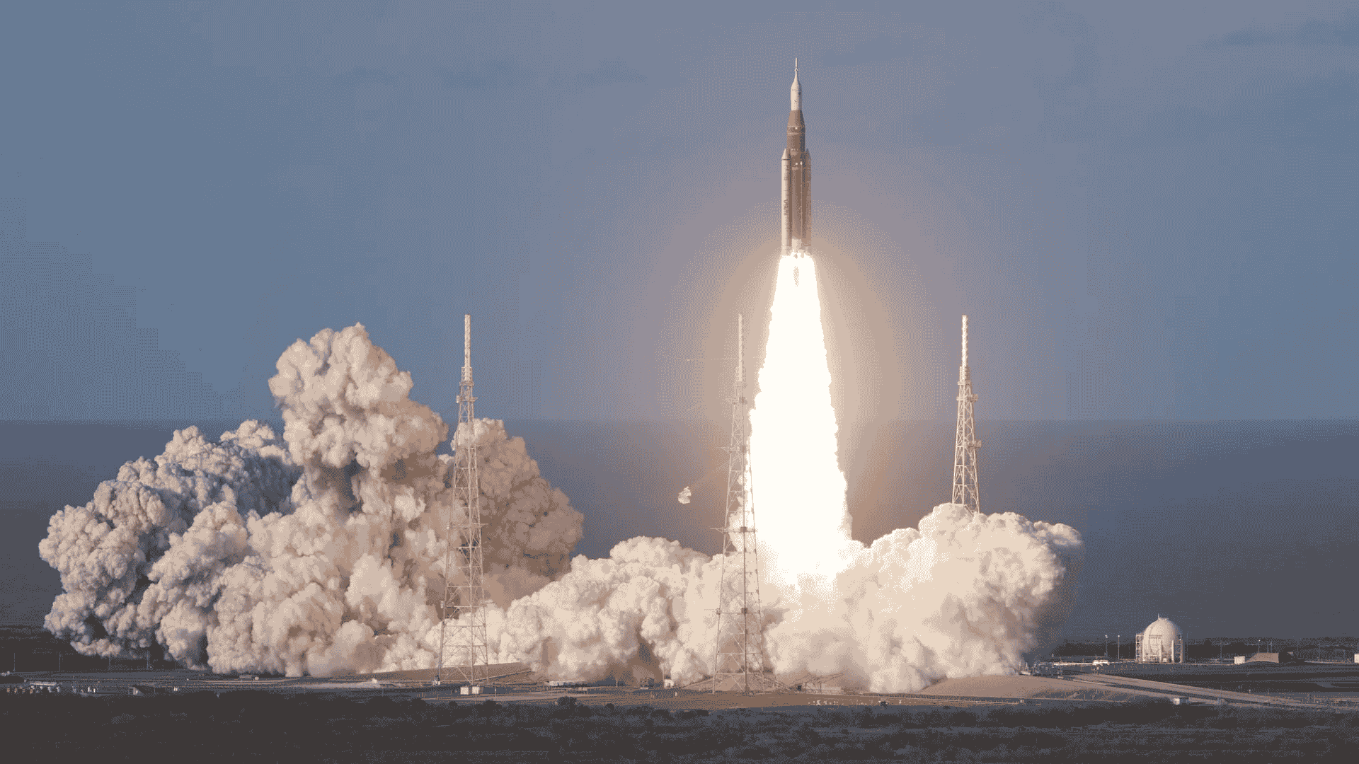 A rocket launches into the sky, surrounded by clouds of smoke and bright flames at its base, reminiscent of the energy at a hot 1027 breakfast, with support towers and a clear sky in the background.