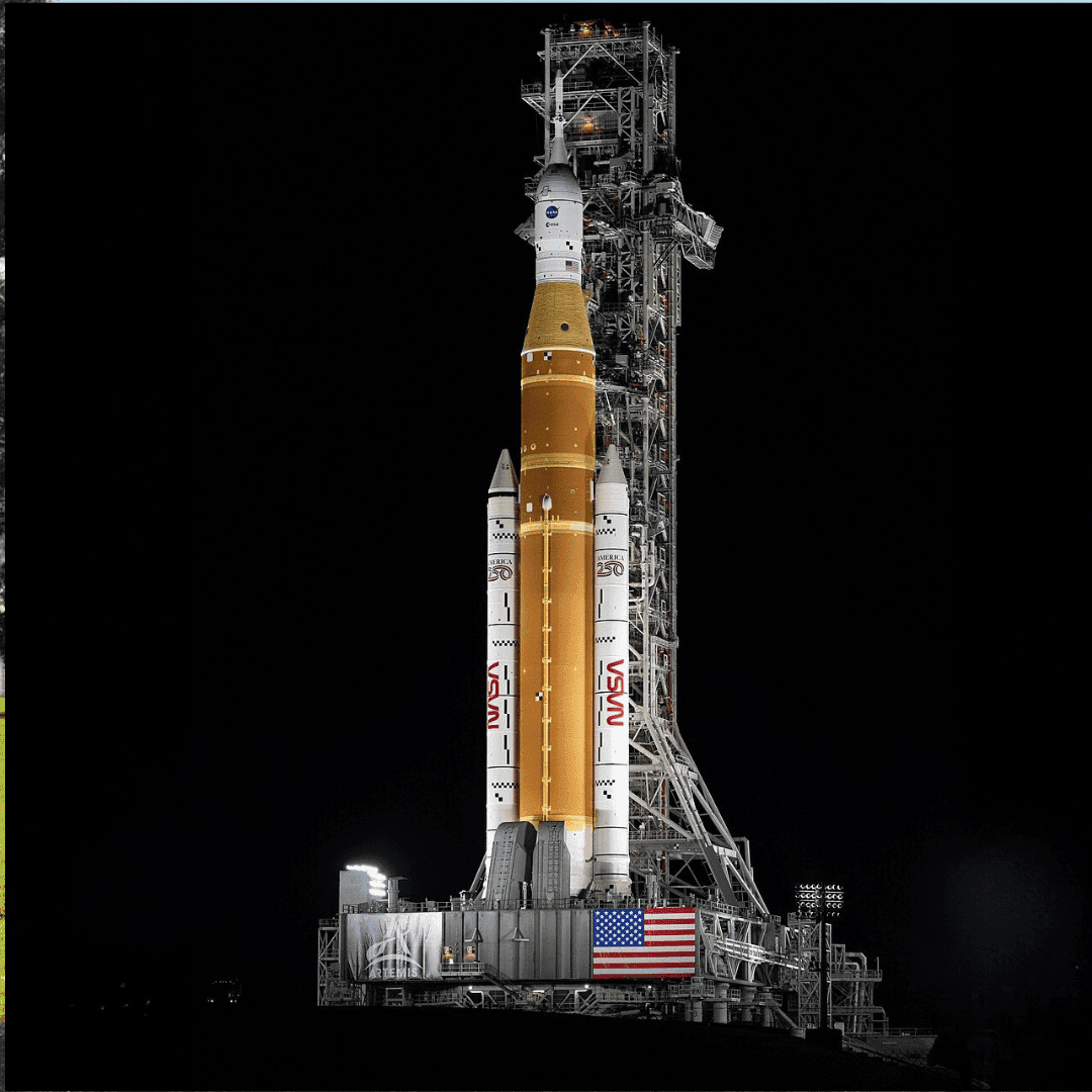 A NASA rocket stands upright on a launch pad at night, illuminated by lights. The orange and white rocket, prepped for fuel relief, is flanked by boosters with a large American flag on the platform and a dark sky behind.