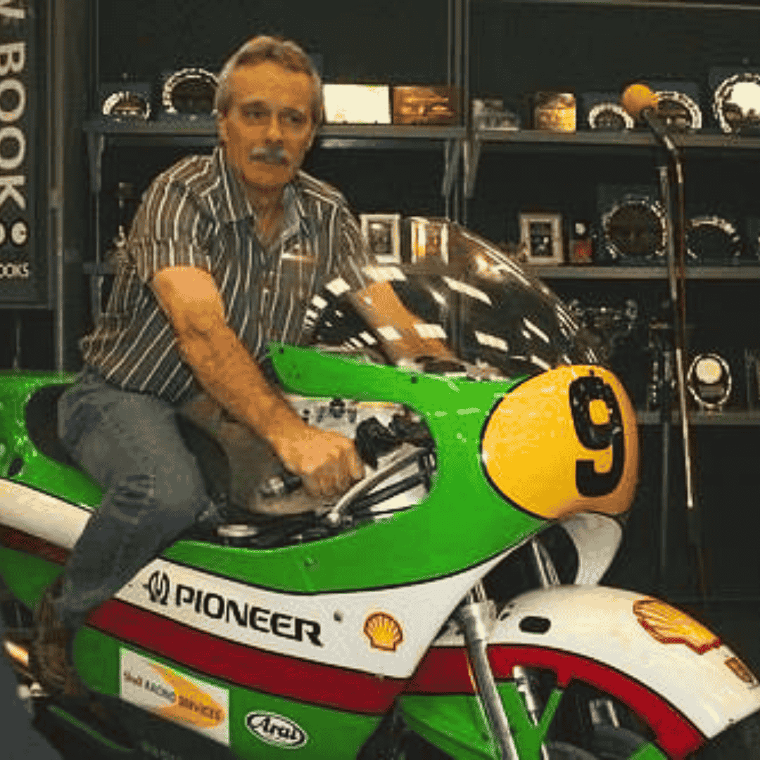 Mark Andrews, a man with gray hair and a mustache, sits on a green and white vintage racing motorcycle numbered 9, indoors and surrounded by shelves filled with plaques and trophies.