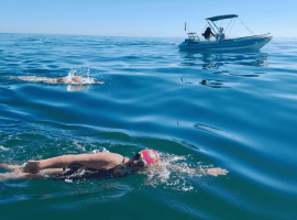 Two swimmers in open water, both wearing swim caps and goggles, swim near a small boat on a calm, sunny day. The boat with people onboard—perhaps filming for Pet TV—follows alongside on the gently rippling blue sea.