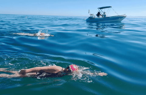 Two swimmers in open water, both wearing swim caps and goggles, swim near a small boat on a calm, sunny day. The boat with people onboard—perhaps filming for Pet TV—follows alongside on the gently rippling blue sea.