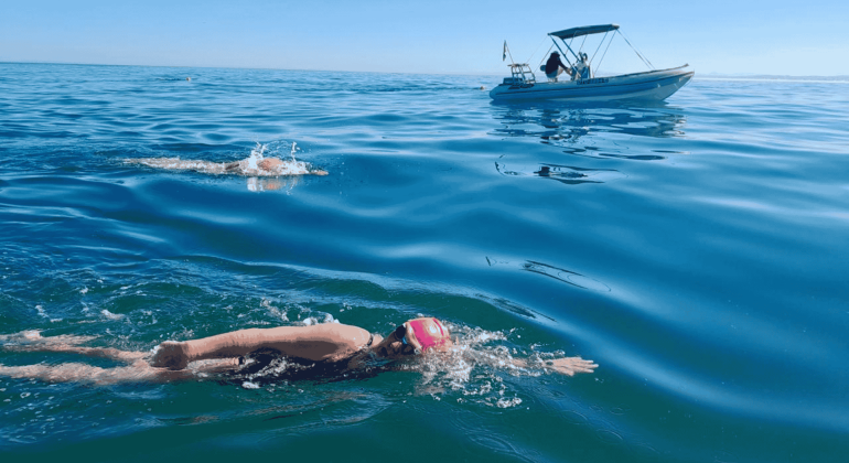 Two swimmers in open water, both wearing swim caps and goggles, swim near a small boat on a calm, sunny day. The boat with people onboard—perhaps filming for Pet TV—follows alongside on the gently rippling blue sea.