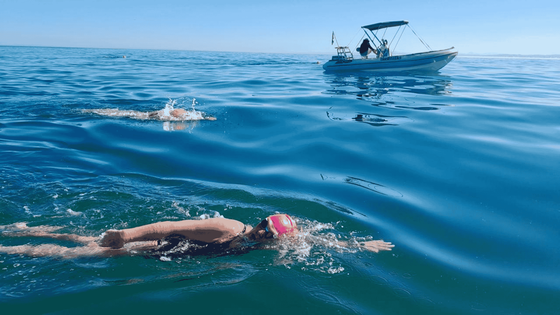 Two swimmers in open water, both wearing swim caps and goggles, swim near a small boat on a calm, sunny day. The boat with people onboard—perhaps filming for Pet TV—follows alongside on the gently rippling blue sea.