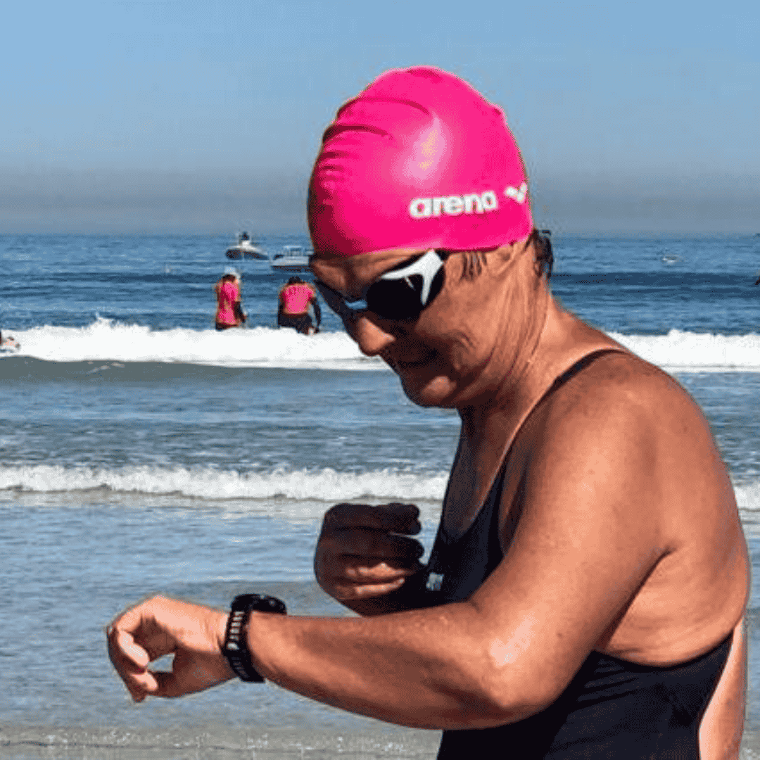 A swimmer in a pink swim cap and goggles checks their watch on a beach, standing near the shoreline. Waves roll in behind them, with others in pink swim caps—capturing a scene as lively as any on Pet TV.