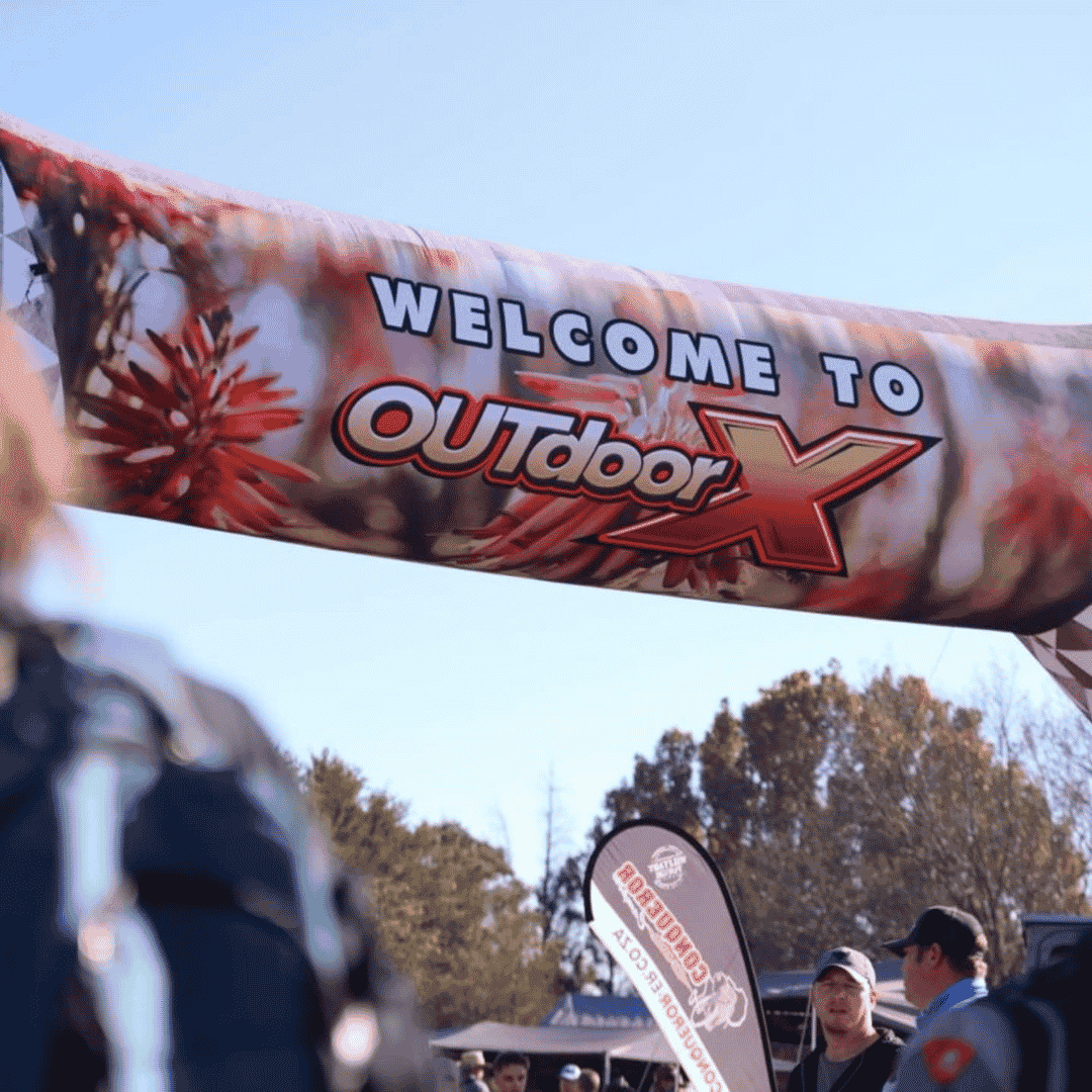 A large banner reading WELCOME TO OUTdoor X hangs above an outdoor event at the Kingsmead Book Fair. People gather below, and trees with autumn leaves are visible in the background under a clear blue sky.